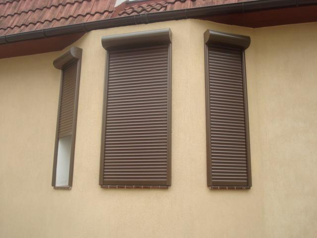 Brązowe rolety zewnętrzne zamontowane na elewacji domu z beżowym tynkiem i czerwoną dachówką, jedna roleta częściowo podniesiona.