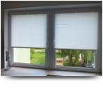 Białe okno PCV z zamontowanymi roletami w kasecie, widok na zielony ogród przez szybę.