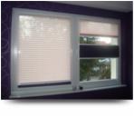 Okno PCV z zamontowanymi białymi plisowanymi osłonami okiennymi, część z nich zasłonięta, widok na zewnątrz przez okno.