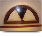 Półokrągłe okno z drewnianą ramą i nietypową żaluzją wewnętrzną w kształcie wachlarza, widoczne z bliska, oświetlone ciepłym światłem.