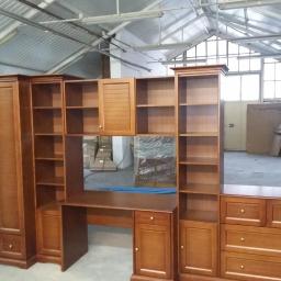 Zakład Stolarski Tadeusz Chodera - Zestaw drewnianych mebli: szafa dwudrzwiowa, regały otwarte, biurko z szafkami i komoda z szufladami, w surowym wnętrzu magazynu.