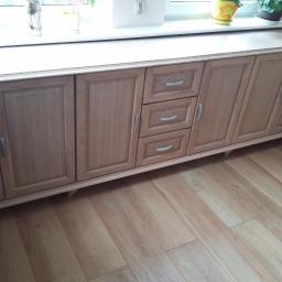 Zakład Stolarski Tadeusz Chodera - Drewniana szafka pod oknem z trzema szufladami i czterema drzwiczkami, wykończona w jasnym odcieniu drewna, z roślinami doniczkowymi na blacie.