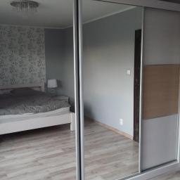 Zakład Stolarski Tadeusz Chodera - Szafa przesuwna z lustrzanymi drzwiami, odbijająca wnętrze sypialni z białym łóżkiem i wzorzystą tapetą.