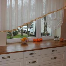Zakład Stolarski Tadeusz Chodera - Fragment kuchni z drewnianym blatem, białymi szafkami z poziomymi uchwytami, owocami w misce i pomarańczami na blacie, widokiem na podwórko z oknem ozdobionym firanką z lamówką.