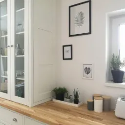 Jasna kuchnia z drewnianym blatem, przeszklona witryna z białymi frontami, na blacie doniczki z roślinami i pojemniki na przyprawy, na ścianie grafiki w czarnych ramkach.