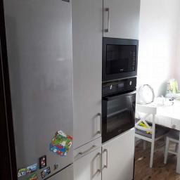 Zakład Stolarski Tadeusz Chodera - Szare meble kuchenne z zabudowaną lodówką, piekarnikiem i mikrofalówką, ozdobione magnesami na lodówce, widok na stół z białymi krzesłami.