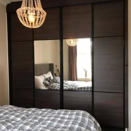 Zakład Stolarski Tadeusz Chodera - Czterodrzwiowa szafa z przesuwnymi drzwiami z ciemnego drewna i lustrzanymi panelami, widoczna w sypialni z łóżkiem w kratę i kryształowym żyrandolem.