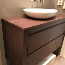 Zakład Stolarski Tadeusz Chodera - Ciemnobrązowa szafka łazienkowa z trzema szufladami i drewnianym blatem, na którym stoi okrągła, biała umywalka nablatowa; w tle widoczna mozaika podłogowa.