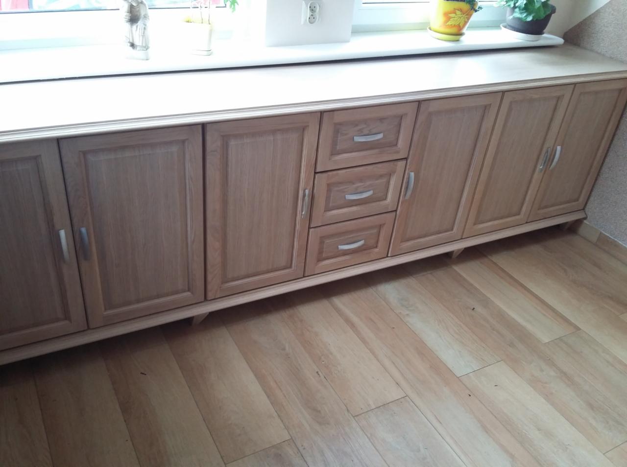 Drewniana szafka pod oknem z trzema szufladami i czterema drzwiczkami, wykończona w jasnym odcieniu drewna, z roślinami doniczkowymi na blacie.