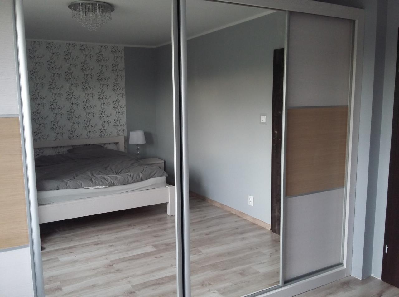 Szafa przesuwna z lustrzanymi drzwiami, odbijająca wnętrze sypialni z białym łóżkiem i wzorzystą tapetą.