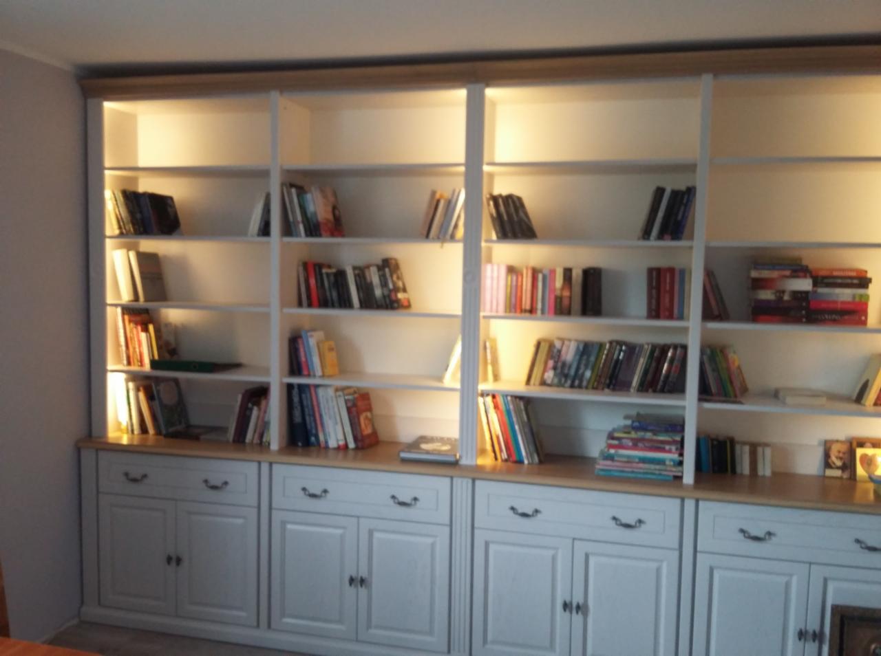 Biała, zabudowana biblioteczka z oświetleniem LED, z dolnymi szafkami z uchwytami muszelkowymi, wypełniona książkami.