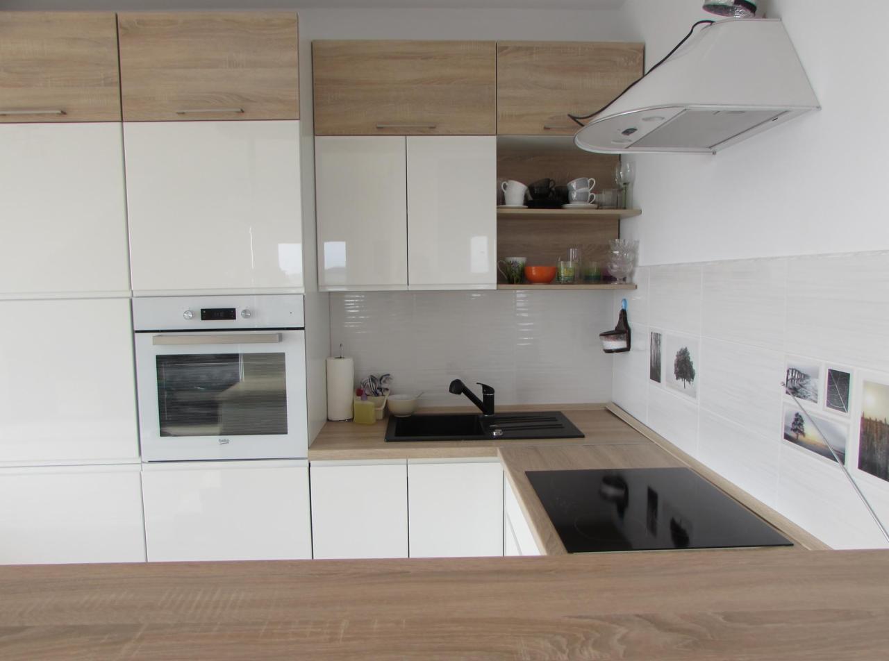 Nowoczesna kuchnia z białymi szafkami dolnymi i górnymi z drewnianymi frontami, czarny zlew i płyta indukcyjna, białe płytki z motywami roślinnymi, drewniany blat na pierwszym planie.