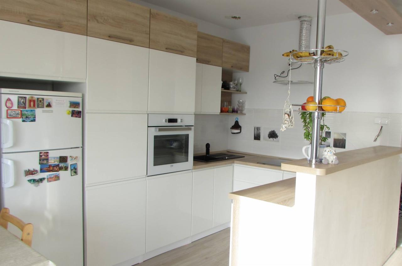 Biała kuchnia na wymiar z drewnianymi akcentami w Sośnie, widok na lodówkę z magnesami, piekarnik i wyspę kuchenną z owocami.