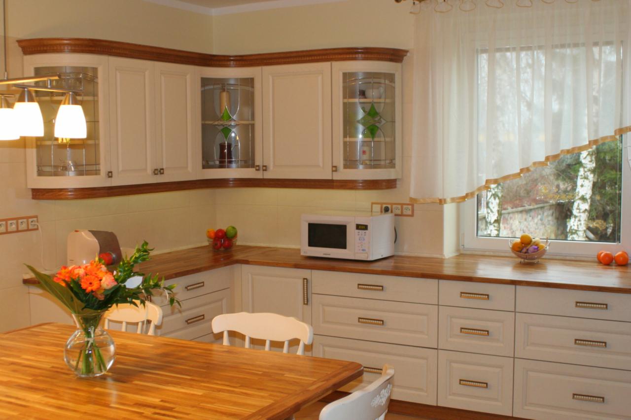 Jasna kuchnia z drewnianymi blatami i frezowanymi frontami, przeszklone szafki wiszące z witrażami, kwiaty w wazonie na stole.