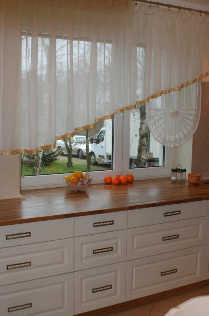 Fragment kuchni z drewnianym blatem, białymi szafkami z poziomymi uchwytami, owocami w misce i pomarańczami na blacie, widokiem na podwórko z oknem ozdobionym firanką z lamówką.