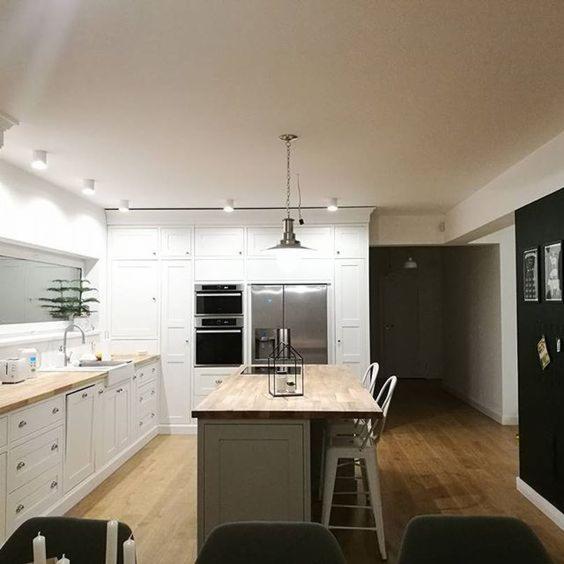 Jasna kuchnia z białymi szafkami, drewnianym blatem i wyspą kuchenną z szarym wykończeniem, oświetlona wiszącą lampą w stylu industrialnym.