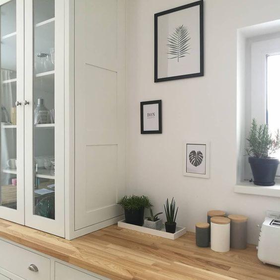 Jasna kuchnia z drewnianym blatem, przeszklona witryna z białymi frontami, na blacie doniczki z roślinami i pojemniki na przyprawy, na ścianie grafiki w czarnych ramkach.