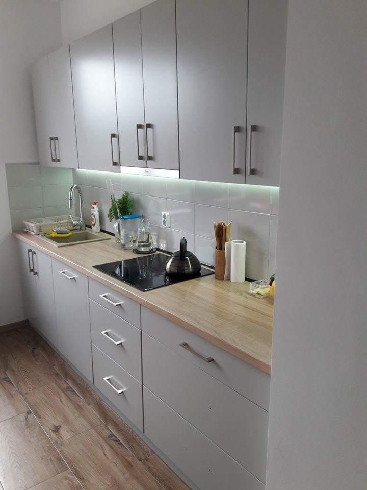 Szara kuchnia z drewnianym blatem, czajnikiem na płycie indukcyjnej, widoczne górne szafki z podświetleniem LED pod spodem i drewniana podłoga.