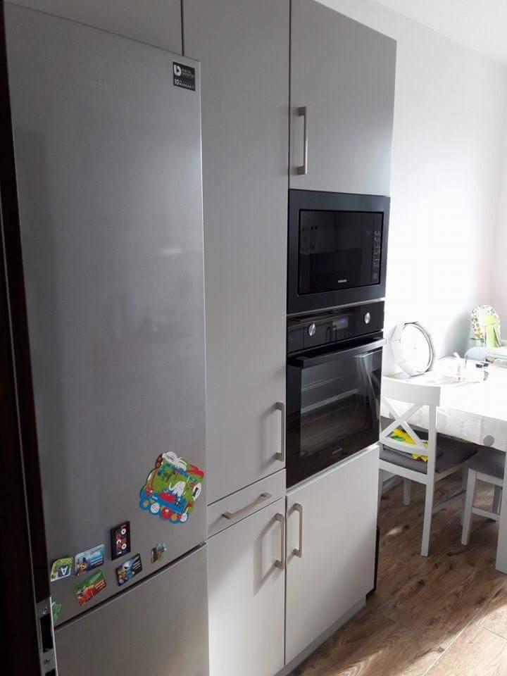 Szare meble kuchenne z zabudowaną lodówką, piekarnikiem i mikrofalówką, ozdobione magnesami na lodówce, widok na stół z białymi krzesłami.