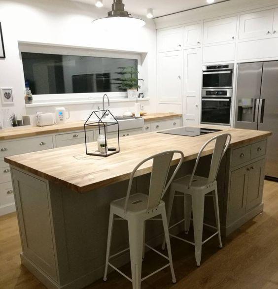 Jasna kuchnia z drewnianym blatem wyspy, dwie białe metalowe hokery i dekoracyjna lampa w kształcie domku na blacie.