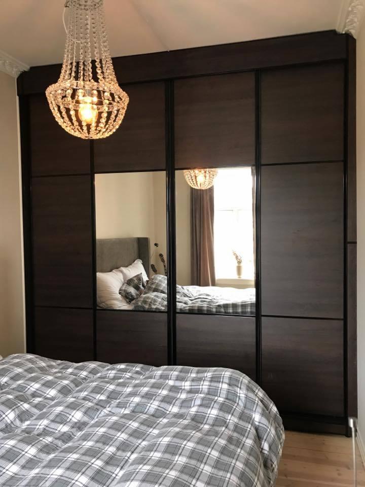Czterodrzwiowa szafa z przesuwnymi drzwiami z ciemnego drewna i lustrzanymi panelami, widoczna w sypialni z łóżkiem w kratę i kryształowym żyrandolem.