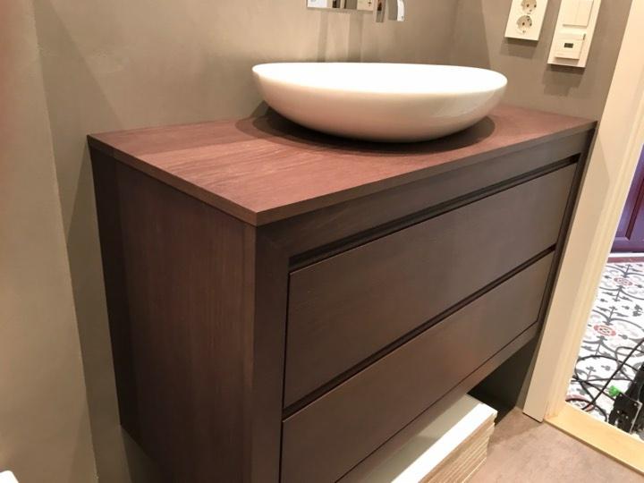 Ciemnobrązowa szafka łazienkowa z trzema szufladami i drewnianym blatem, na którym stoi okrągła, biała umywalka nablatowa; w tle widoczna mozaika podłogowa.