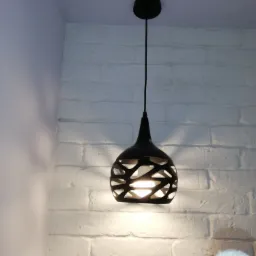 Czarna, ażurowa lampa wisząca o stożkowym kształcie na tle ściany z białej cegły, oświetlona ciepłym światłem żarówki.