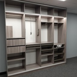 CHISEL KUCHNIE Agnieszka Kowalicka - Wnętrze szafy z szarego drewna z oświetleniem LED, z wysuwanymi szufladami na akcesoria, drążkami na ubrania i półkami, na tle szarej ściany i ciemnej wykładziny.