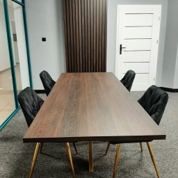 CHISEL KUCHNIE Agnieszka Kowalicka - Stół z blatem drewnianym o ciemnym odcieniu, ustawiony w biurze z czterema krzesłami tapicerowanymi i złotymi nogami. W tle ściana z drewnianymi panelami oraz białe drzwi.