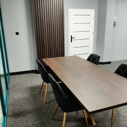 CHISEL KUCHNIE Agnieszka Kowalicka - Stół z blatem drewnianym w minimalistycznym wnętrzu biurowym z czarnymi krzesłami na złotych nogach i dekoracyjną ścianą z drewnianych listew.