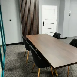 Stół z blatem drewnianym w minimalistycznym wnętrzu biurowym z czarnymi krzesłami na złotych nogach i dekoracyjną ścianą z drewnianych listew.