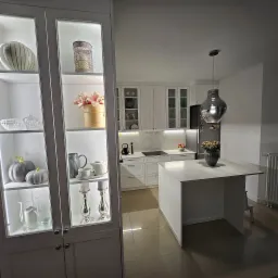 Nowoczesna biała kuchnia z wyspą, oświetlonymi witrynami z dekoracjami i designerską lampą wiszącą.