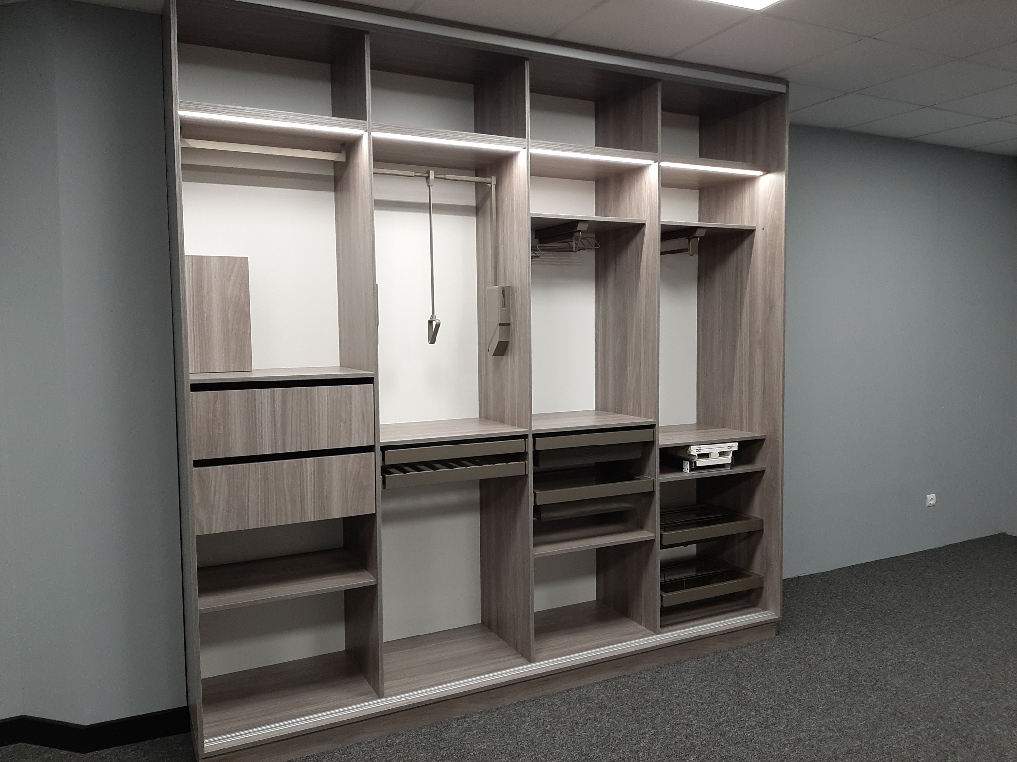 Wnętrze szafy z szarego drewna z oświetleniem LED, z wysuwanymi szufladami na akcesoria, drążkami na ubrania i półkami, na tle szarej ściany i ciemnej wykładziny.