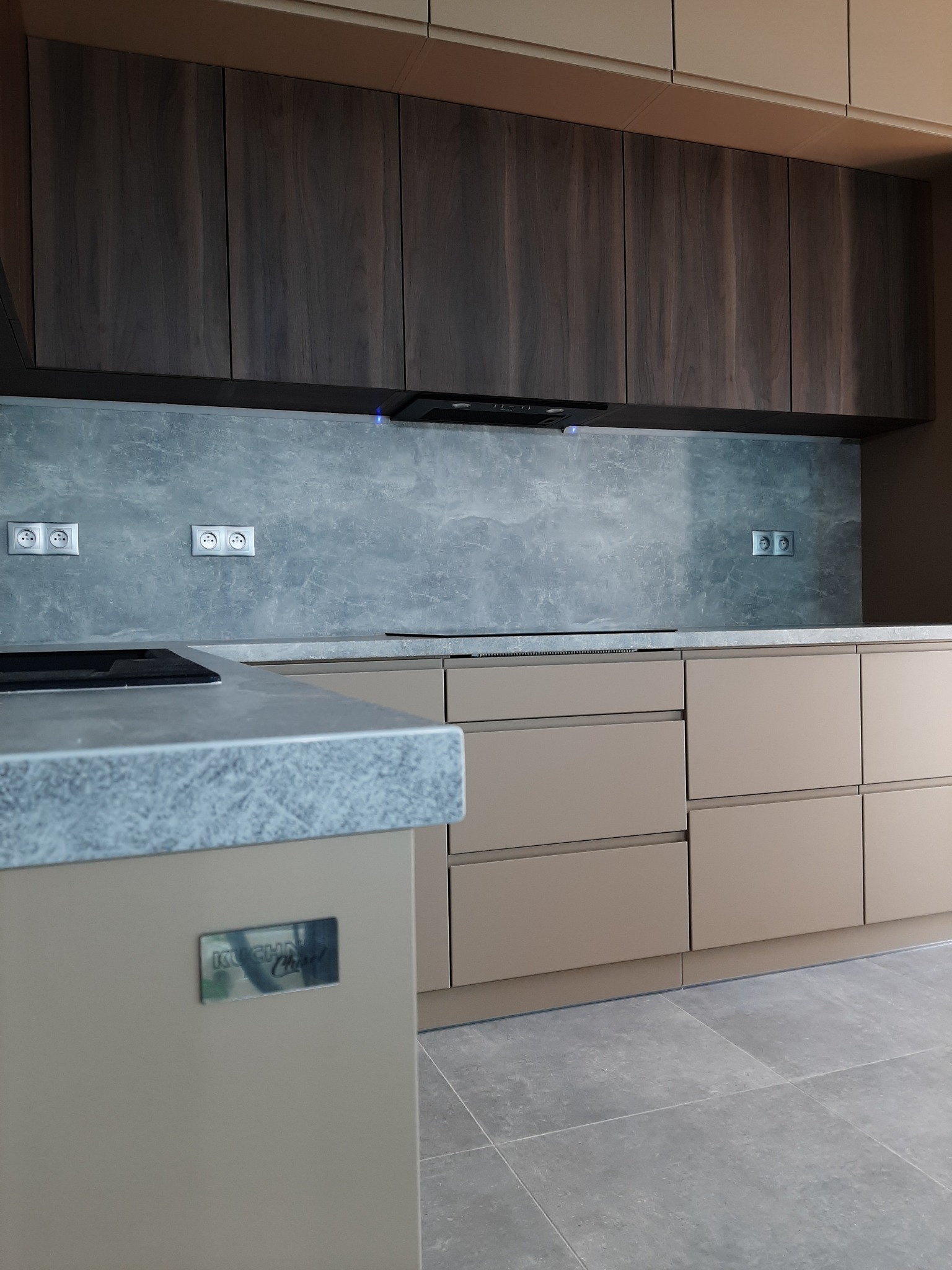 Nowoczesna kuchnia w Legnicy z beżowymi szafkami dolnymi i ciemnymi, drewnopodobnymi szafkami górnymi, szarym blatem i marmurową ścianą nad blatem, z widocznym fragmentem płyty indukcyjnej.