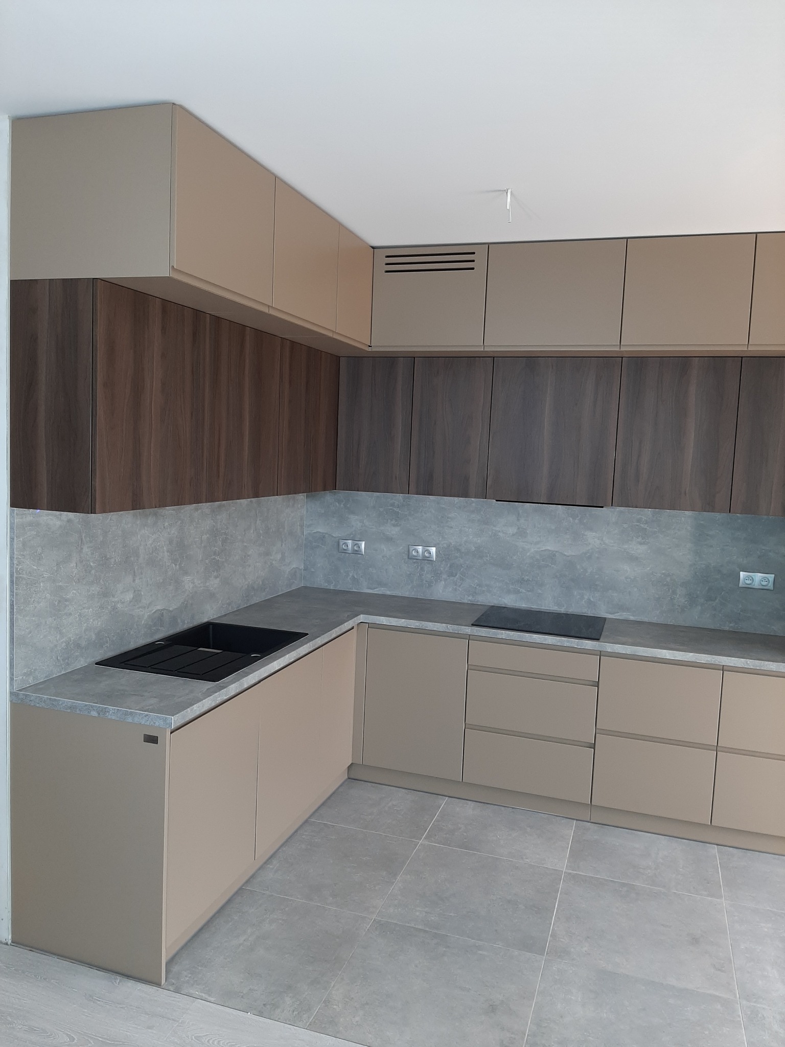 Nowoczesna kuchnia z beżowymi frontami szafek dolnych i górnych, ciemnymi, drewnopodobnymi szafkami górnymi, szarym blatem i backsplashiem imitującym kamień, czarnym zlewem i płytą indukcyjną.