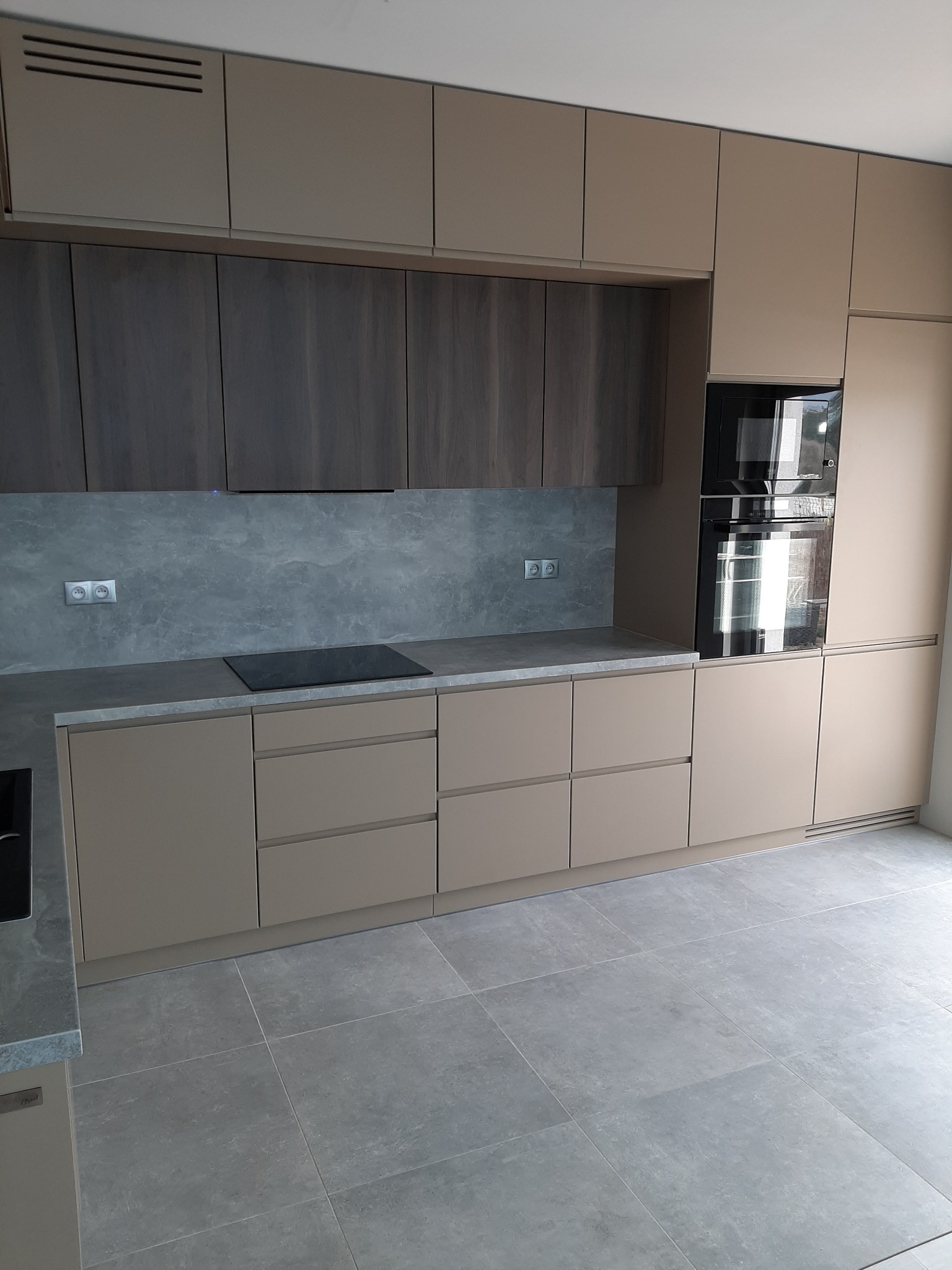 Nowoczesna kuchnia: beżowe i grafitowe szafki z drewnianym fornirem, szary blat i backsplash imitujący kamień, płyta indukcyjna, zabudowane piekarniki.