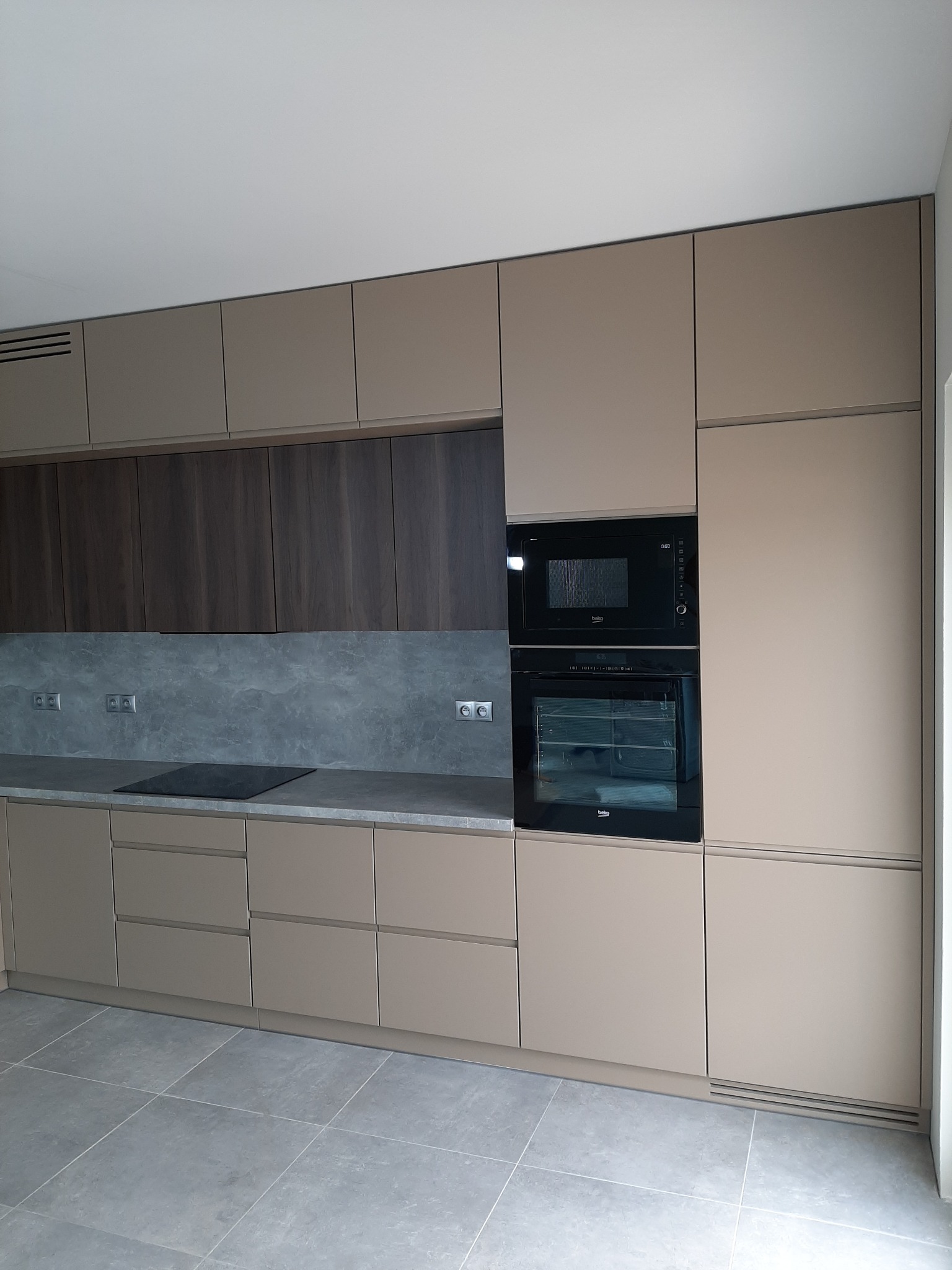 Nowoczesna kuchnia z zabudową w odcieniach beżu i brązu, z wbudowanym piekarnikiem i płytą indukcyjną na szarym blacie. Minimalistyczny design, gładkie fronty szafek bez uchwytów.