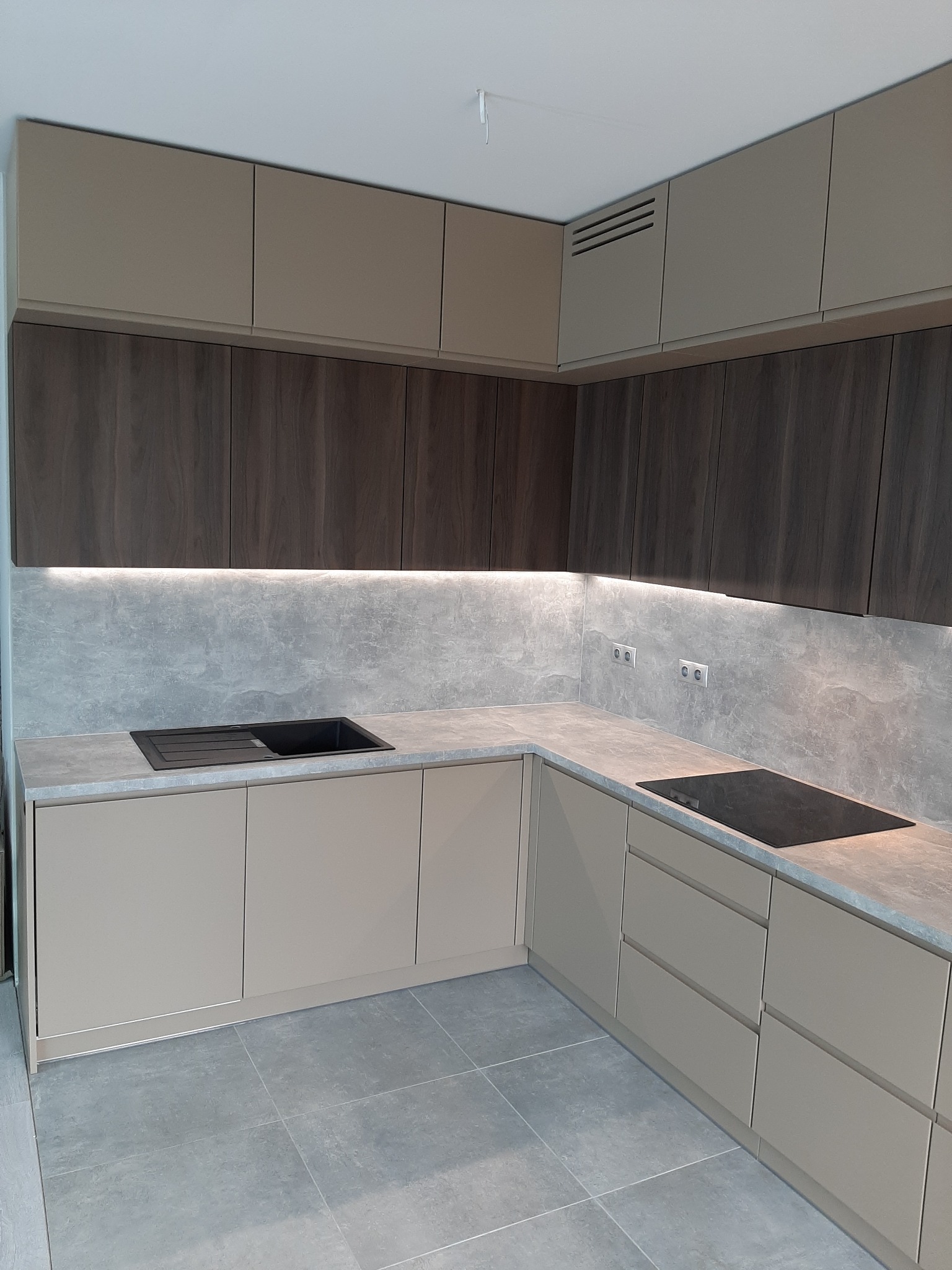 Nowoczesna kuchnia na wymiar z szarymi płytkami podłogowymi, beżowymi dolnymi szafkami i górnymi szafkami w kolorze drewna, podświetlona listwą LED pod szafkami wiszącymi.