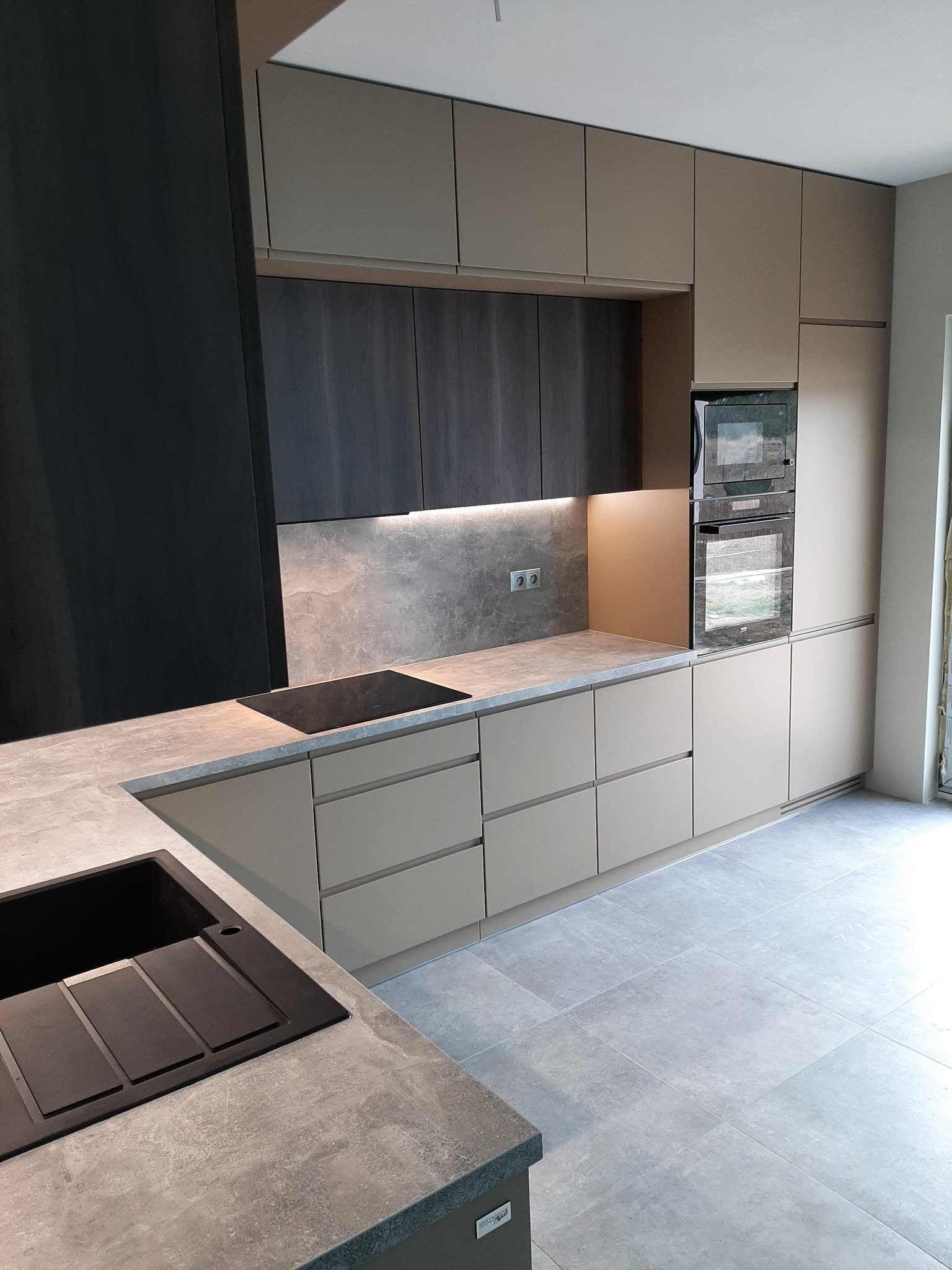 Nowoczesna kuchnia z beżowymi szafkami, ciemnoszarymi elementami dekoracyjnymi, blatem imitującym beton i czarnym zlewem, oświetlona listwą LED pod szafkami wiszącymi.