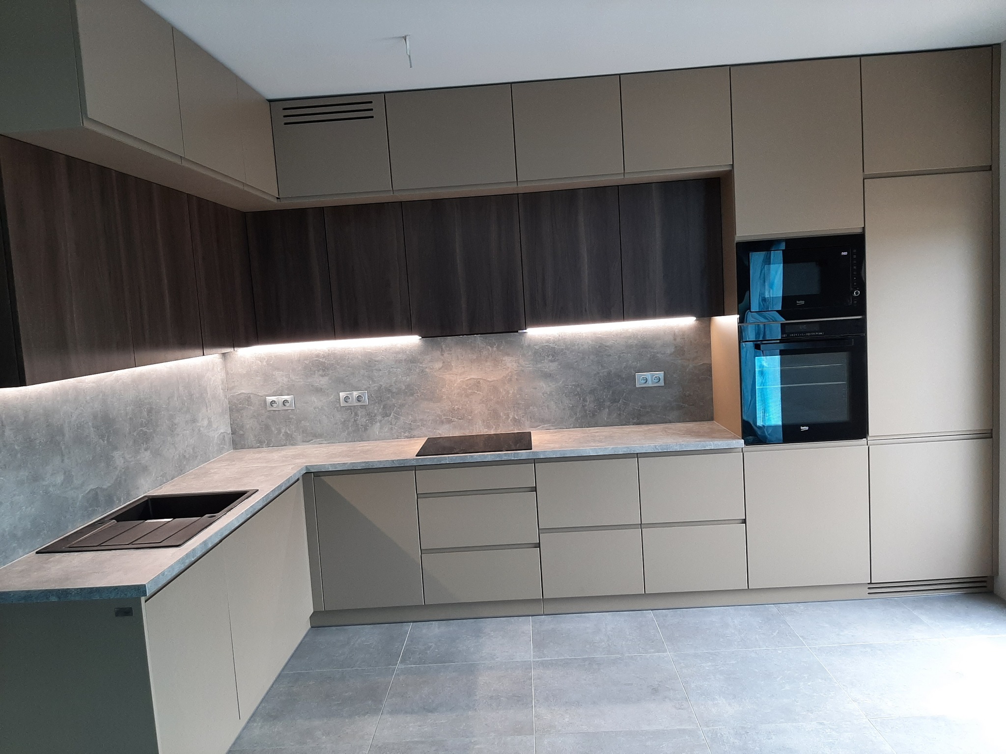 Nowoczesna kuchnia z zabudową w odcieniach beżu i grafitu, oświetlenie LED pod szafkami wiszącymi, płyta indukcyjna na blacie z szarego konglomeratu, piekarnik w zabudowie.