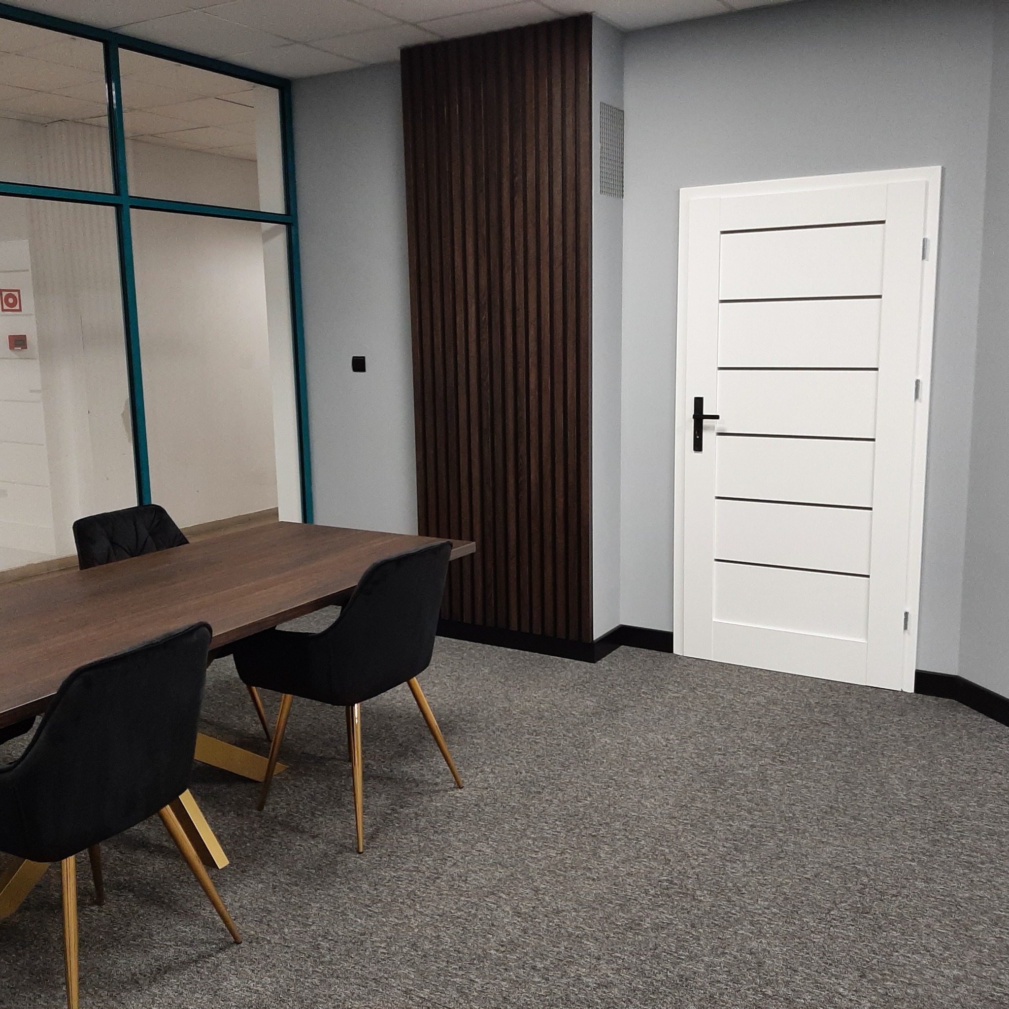 Nowoczesna sala konferencyjna z drewnianym stołem, czarnymi krzesłami na złotych nogach, panelem ściennym z ciemnego drewna i białą, minimalistyczną ścianą działową z przeszkleniami.