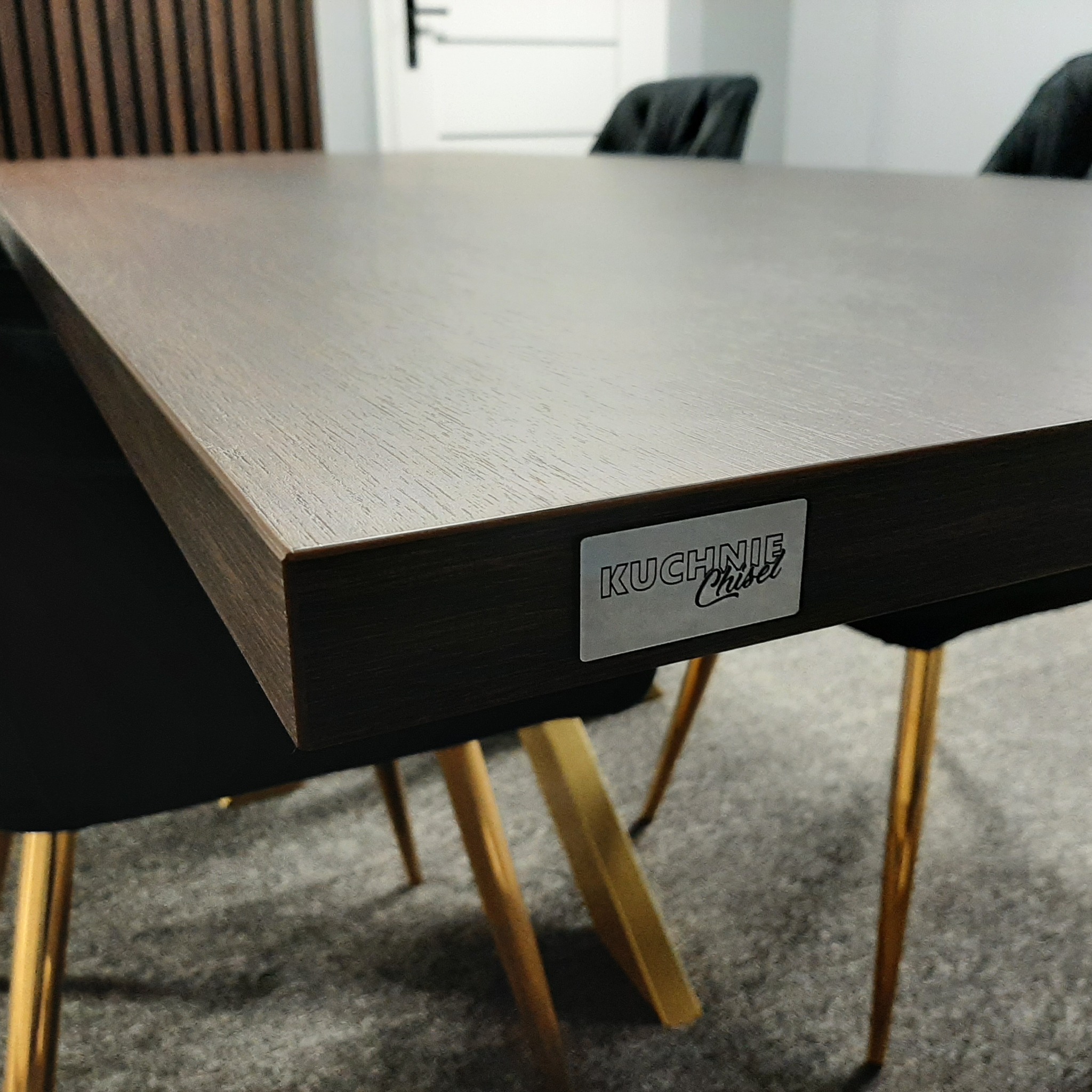 Brązowy blat stołu z widocznym logo 'KUCHNIE Chisel', ustawiony na krzesłach z czarnymi siedziskami i złotymi nogami.