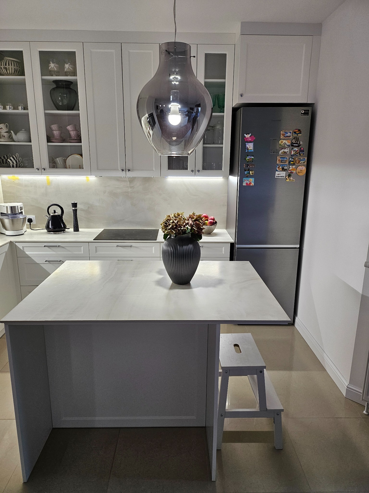 Nowoczesna kuchnia z białymi szafkami, wyspą z blatem imitującym marmur, szklanymi witrynami i srebrną lodówką ozdobioną magnesami, oświetlona wiszącą lampą o dymionym szkle.