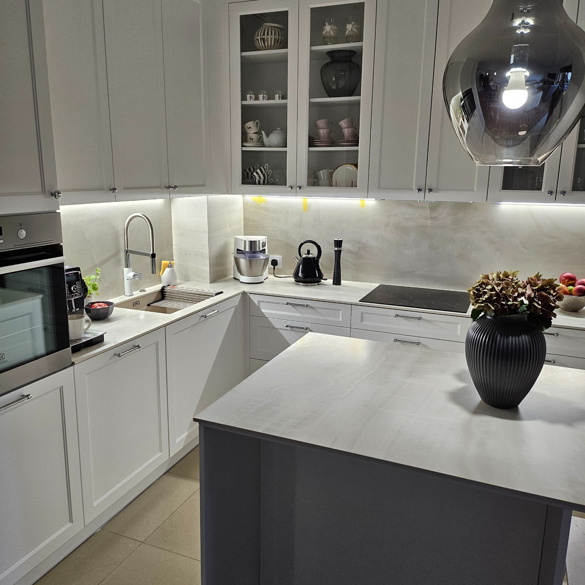 Jasna kuchnia z wyspą, przeszklone szafki górne eksponują dekoracyjną ceramikę, na blacie czajnik elektryczny i robot kuchenny, nad wyspą lampa w kształcie kropli.