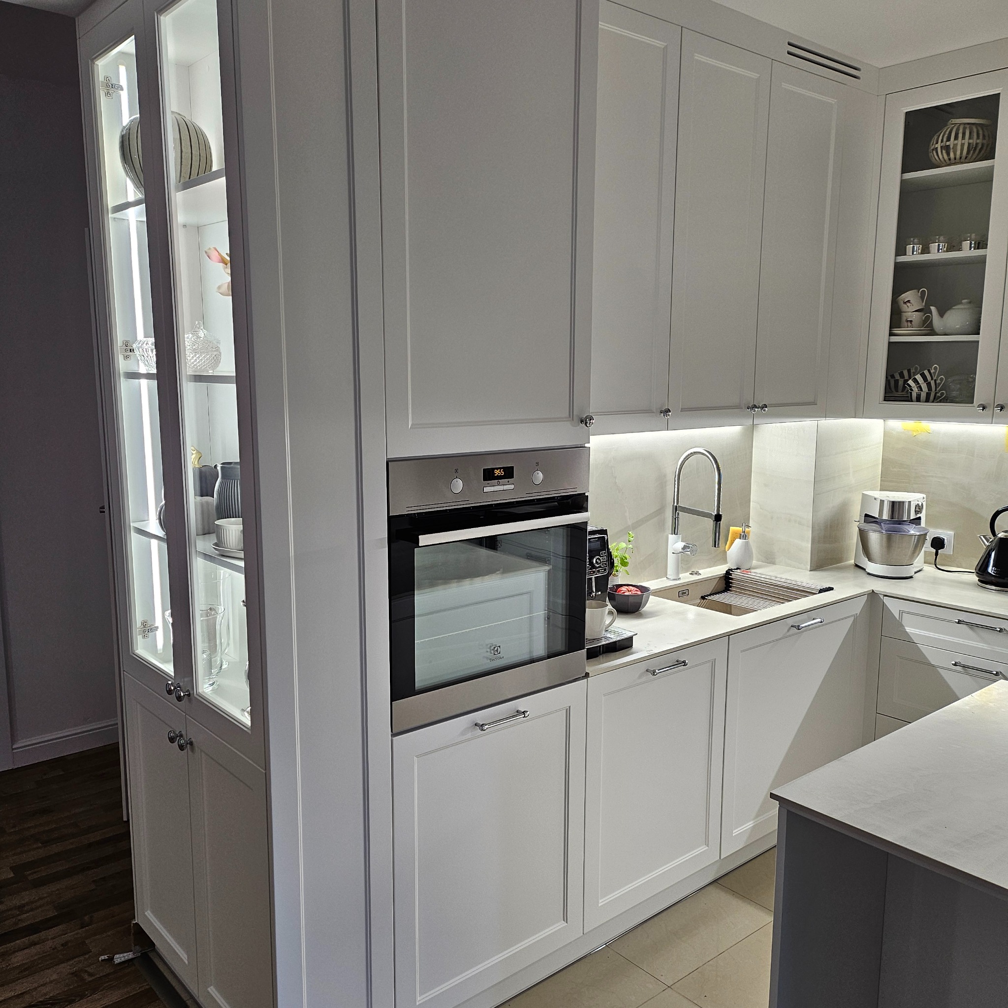 Jasna kuchnia z białymi, frezowanymi frontami, szklaną witryną z podświetleniem LED, piekarnikiem Electrolux i blatem z kamienia kompozytowego.
