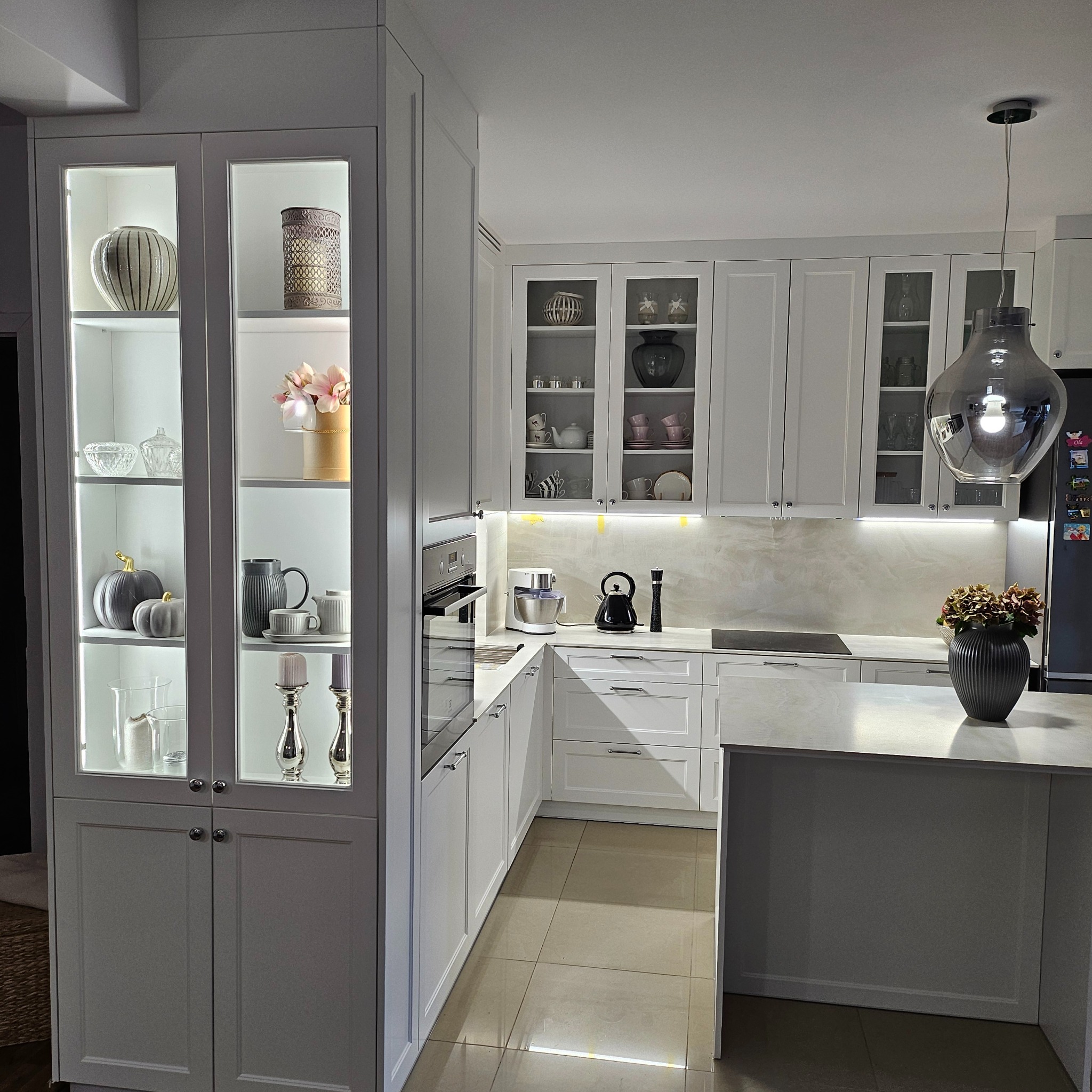 Jasna, nowoczesna kuchnia z wyspą i przeszkloną witryną w Legnicy, oświetlona listwami LED, z dekoracyjnymi wazonami i naczyniami wewnątrz.