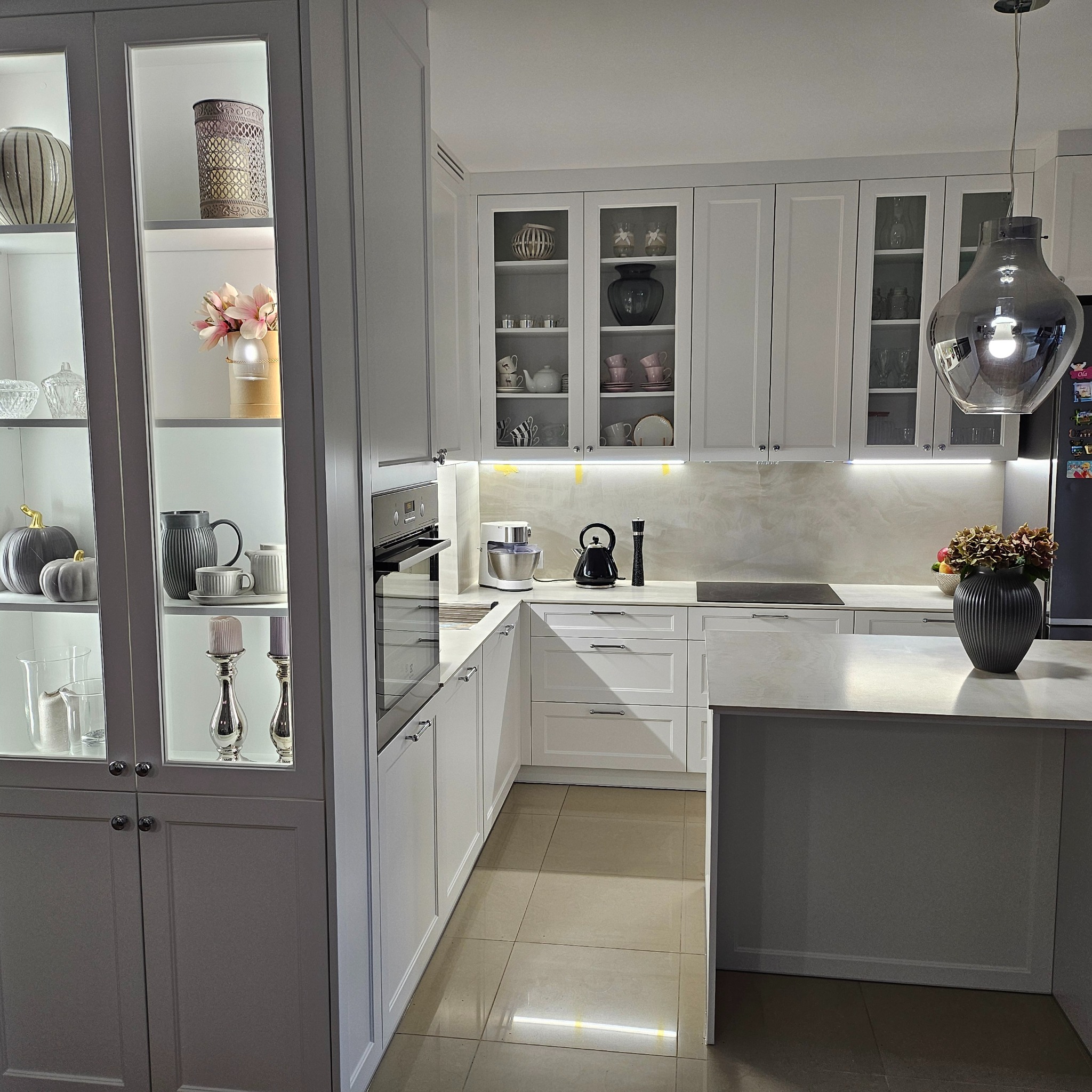 Nowoczesna, jasna kuchnia z białymi frontami, przeszklonymi szafkami, wyspą i dekoracyjnymi elementami, takimi jak wazony i owoce, oświetlona wiszącą lampą.