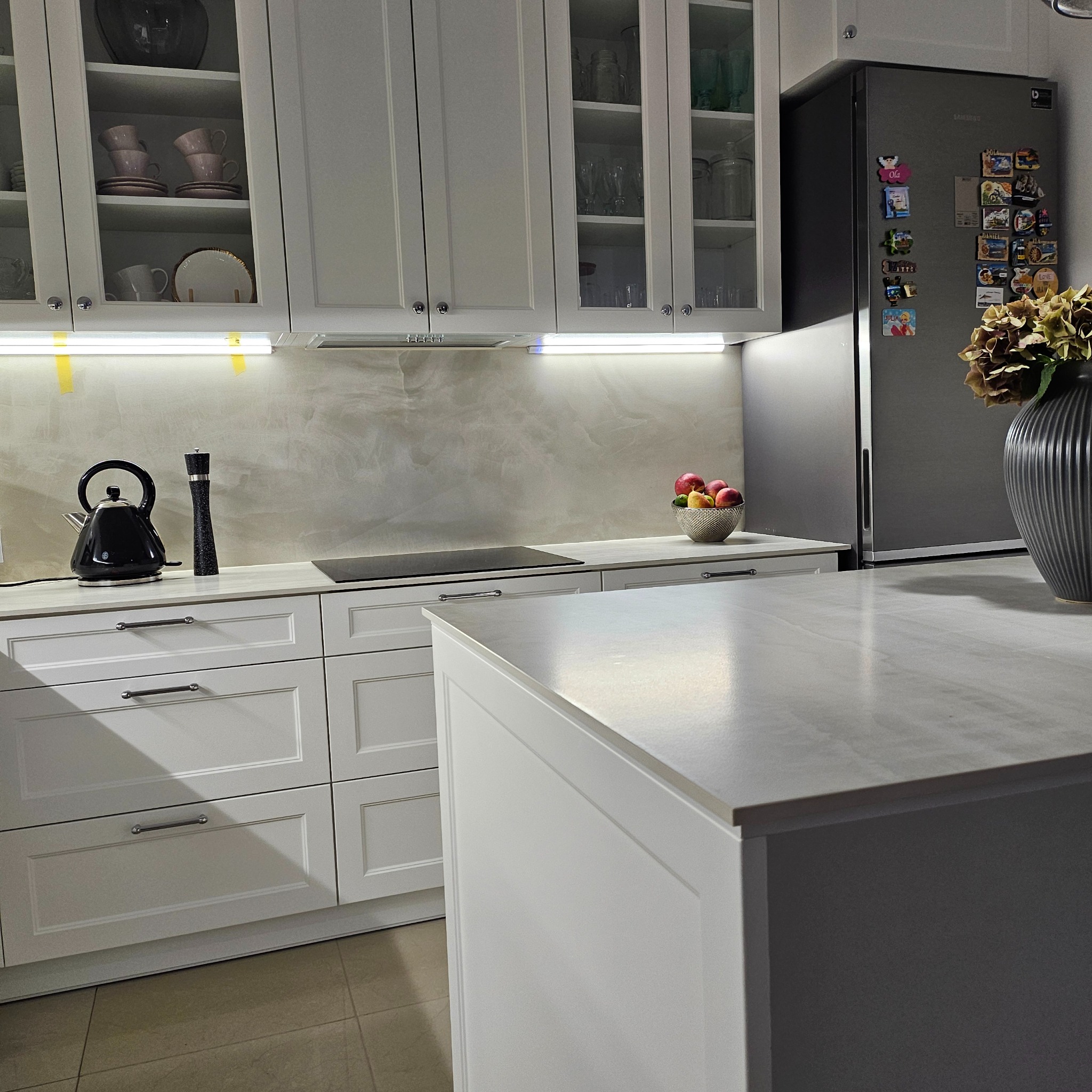 Nowoczesna, jasna kuchnia z białymi frontami szafek, szklanymi witrynami, wyspą kuchenną i szarym blatem. Na blacie czajnik, młynek do pieprzu i misa z owocami. Lodówka obok z magnesami.