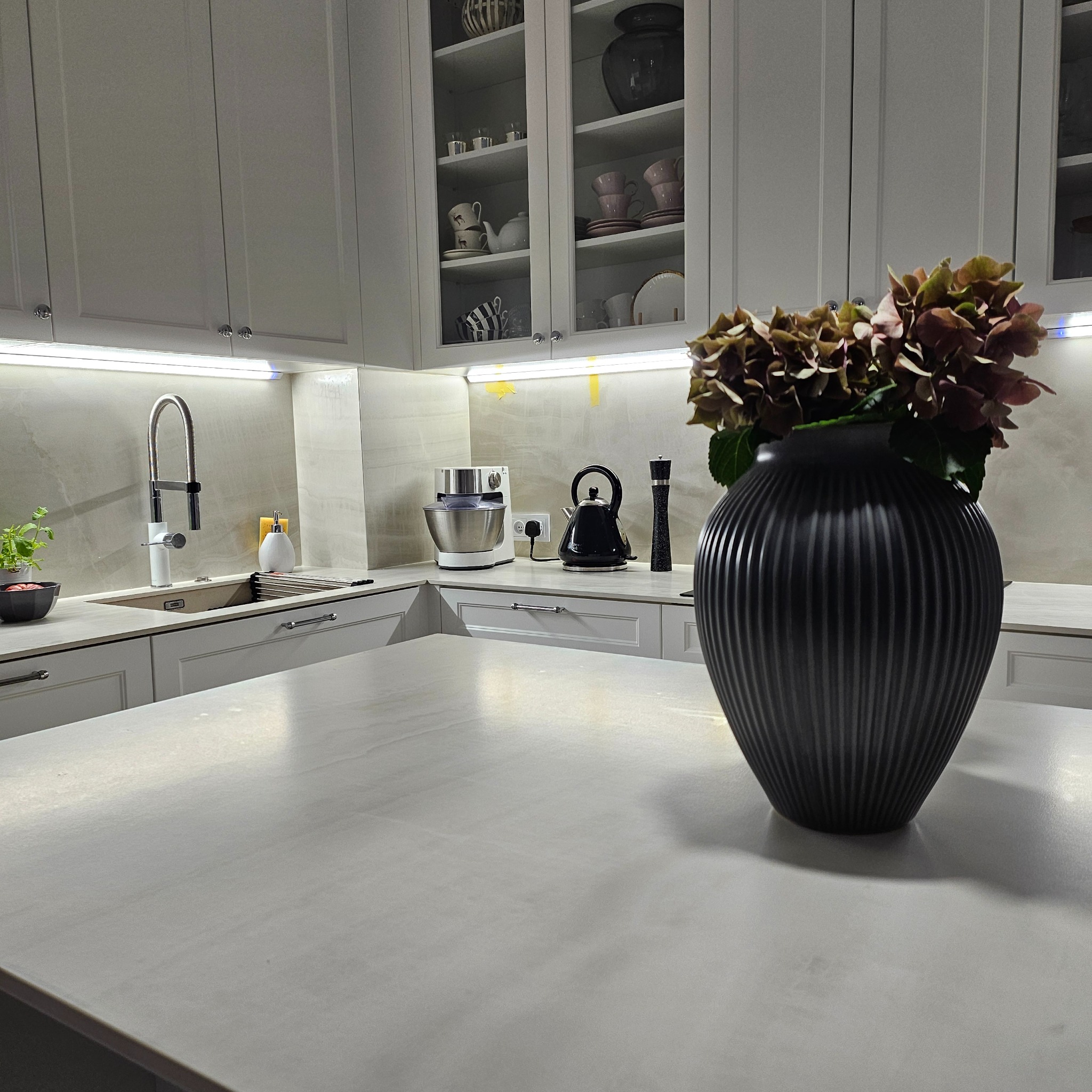 Nowoczesna, jasna kuchnia z białymi szafkami i blatem w odcieniach szarości, oświetlona listwami LED, z czarnym wazonem z hortensjami na pierwszym planie.