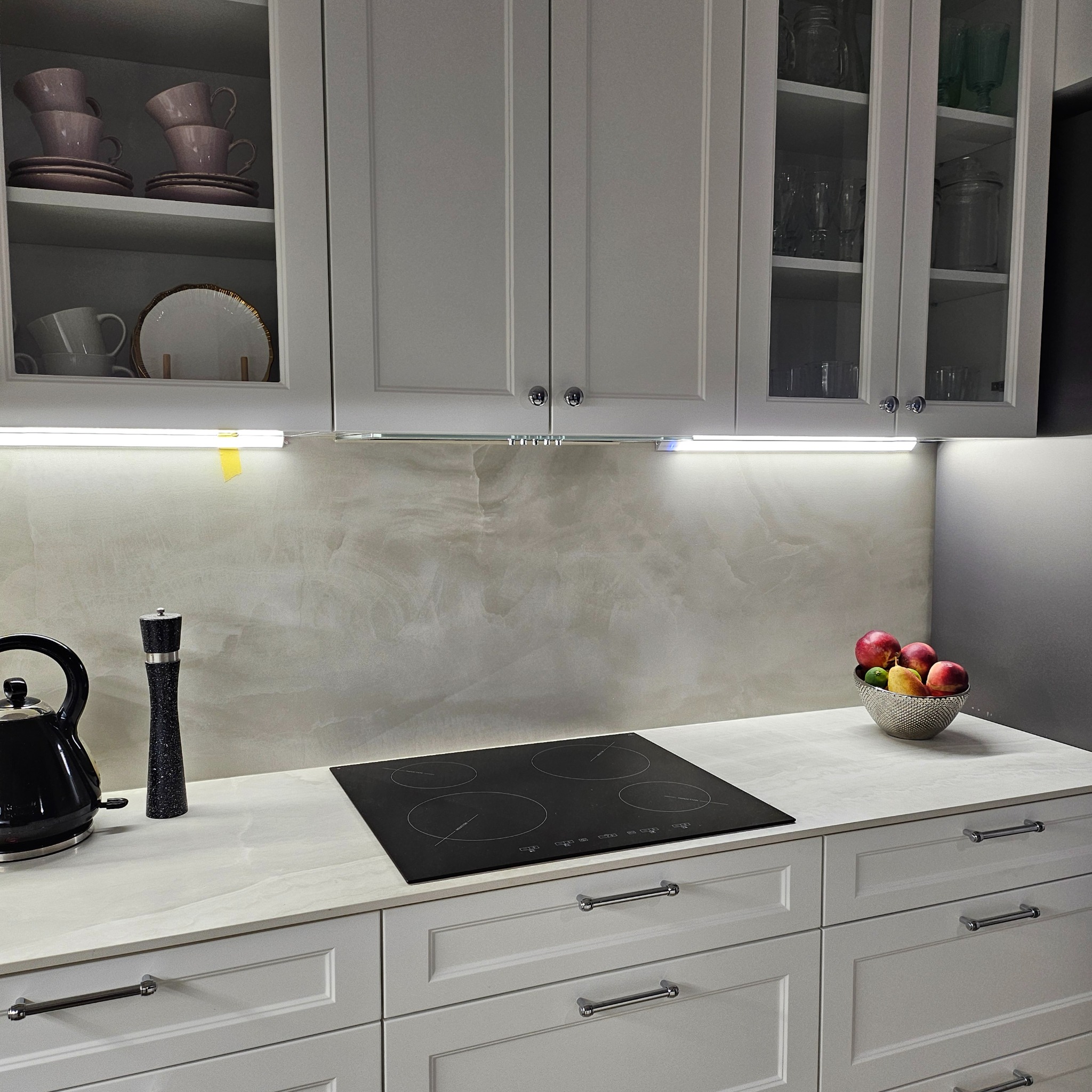 Jasnoszara kuchnia na wymiar z białym blatem, czarną płytą indukcyjną, czajnikiem, młynkiem do pieprzu i owocami w metalowej misie, oświetlona listwą LED pod szafkami wiszącymi.